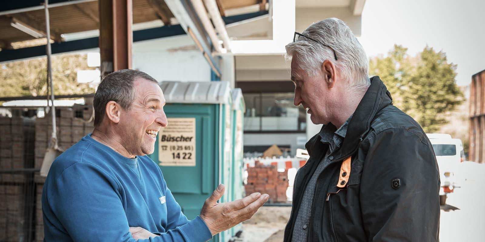 Ein lächelnder Mann in einem blauen Pullover gestikuliert lebhaft, während sein Gesprächspartner aufmerksam zuhört.