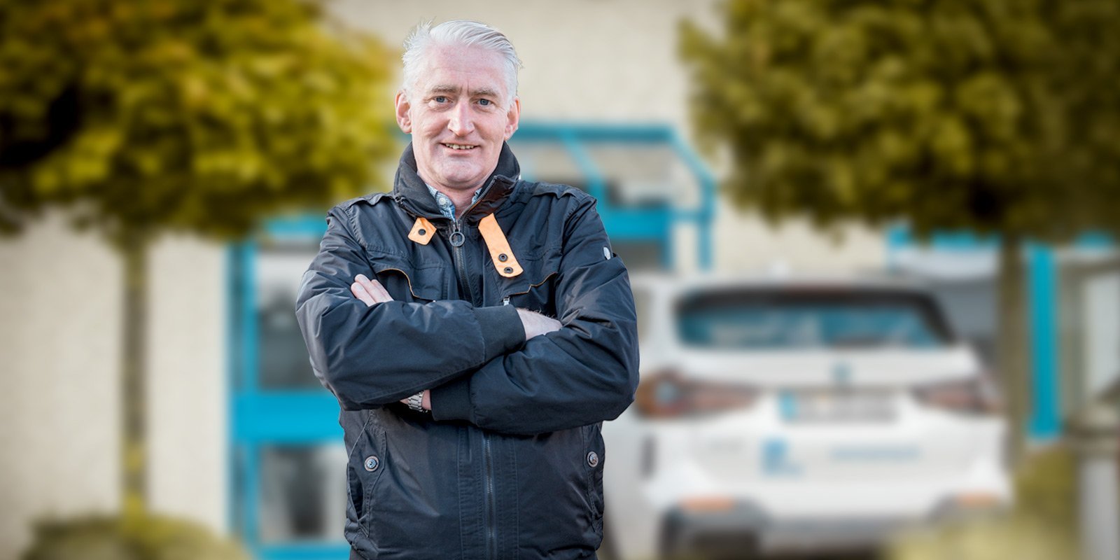 Ein Mann mit grauen Haaren steht lächelnd mit verschränkten Armen vor einem Gebäude, umgeben von grünen Bäumen.